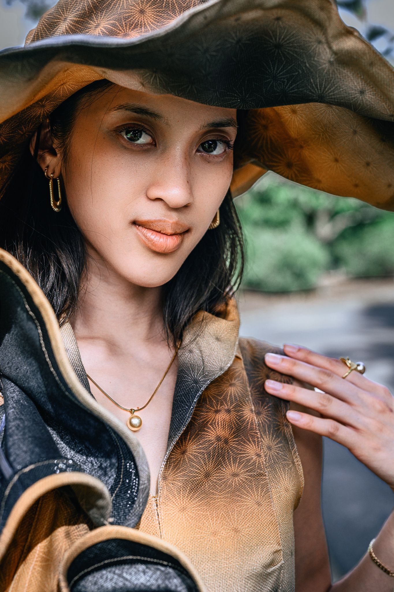 Young woman in wide-brim hat, gold necklace, and leather jacket poses outdoors in a stylish boho-chic portrait.