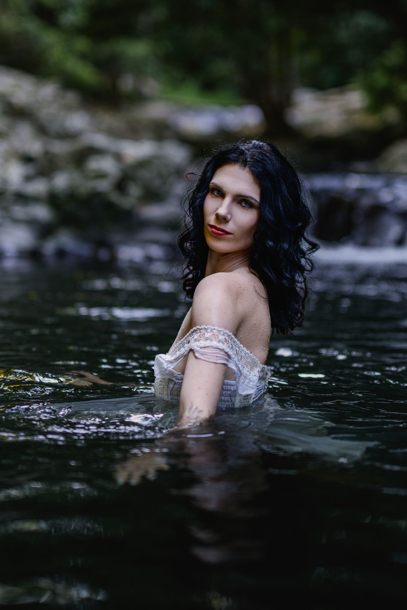 Dark-haired woman in white off-shoulder dress standing in a forest stream, gazing at camera.