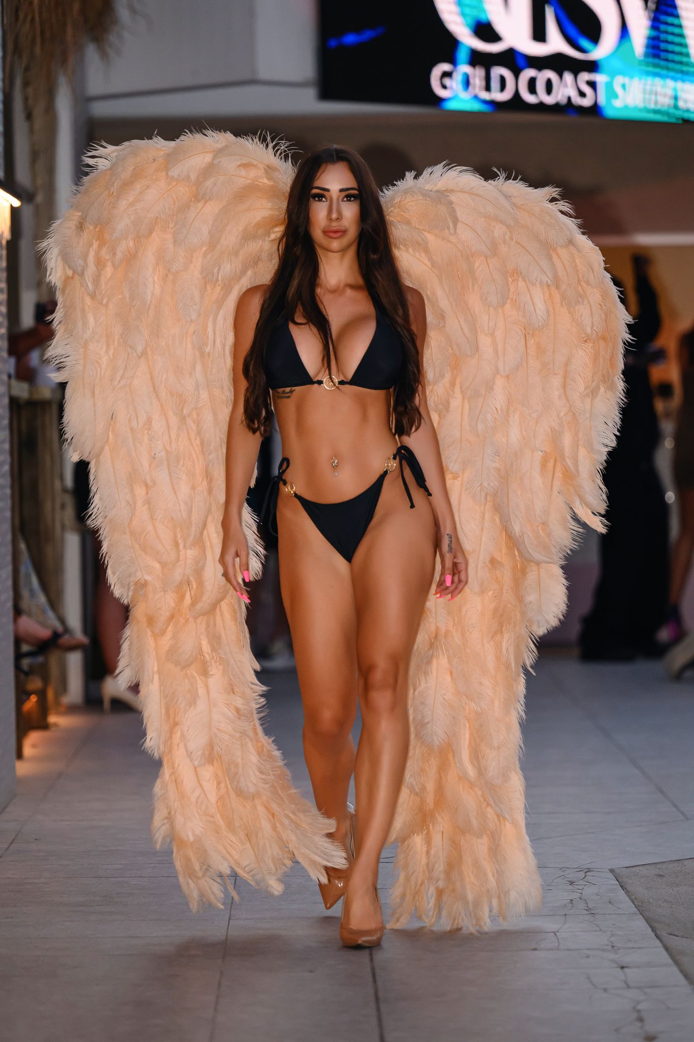 Model presents black swimwear with peach-colored feathered wing accessories on catwalk.