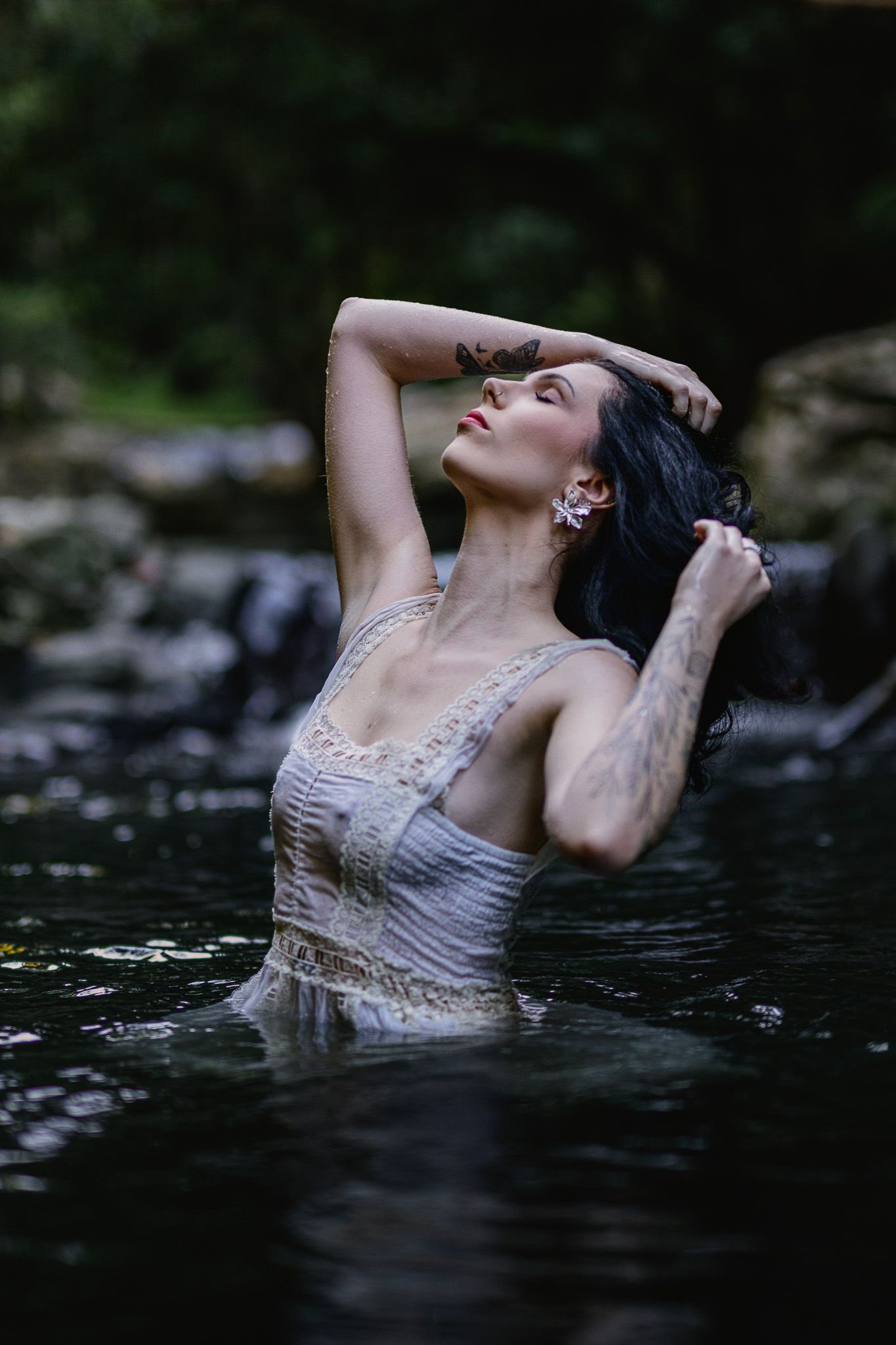 A tattooed woman in a white lace dress stands waist-deep in a serene forest river, posing gracefully with arms raised.