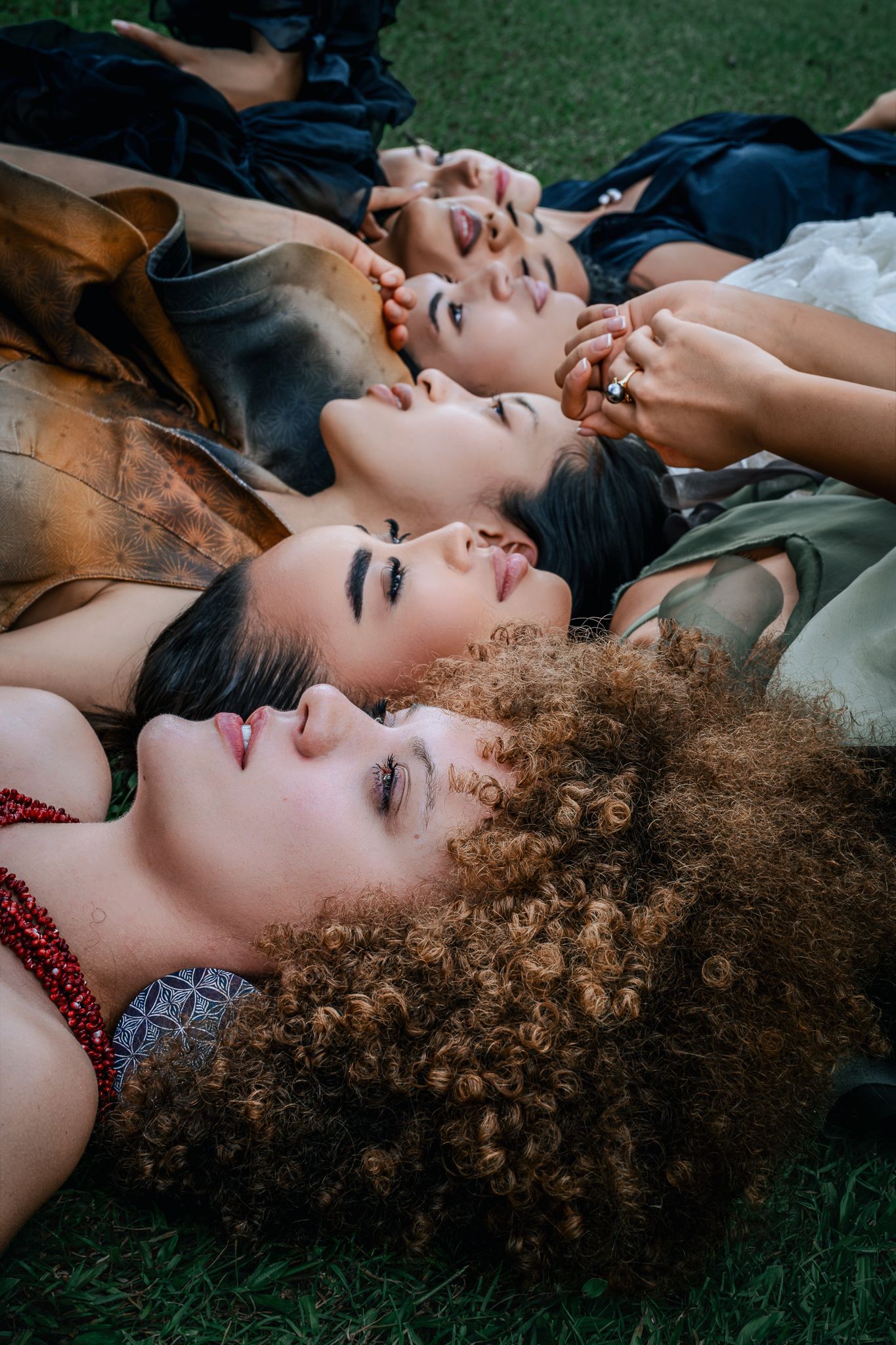 Five diverse women lying on grass in elegant dresses, showcasing natural beauty and unity in a stylish outdoor portrait.