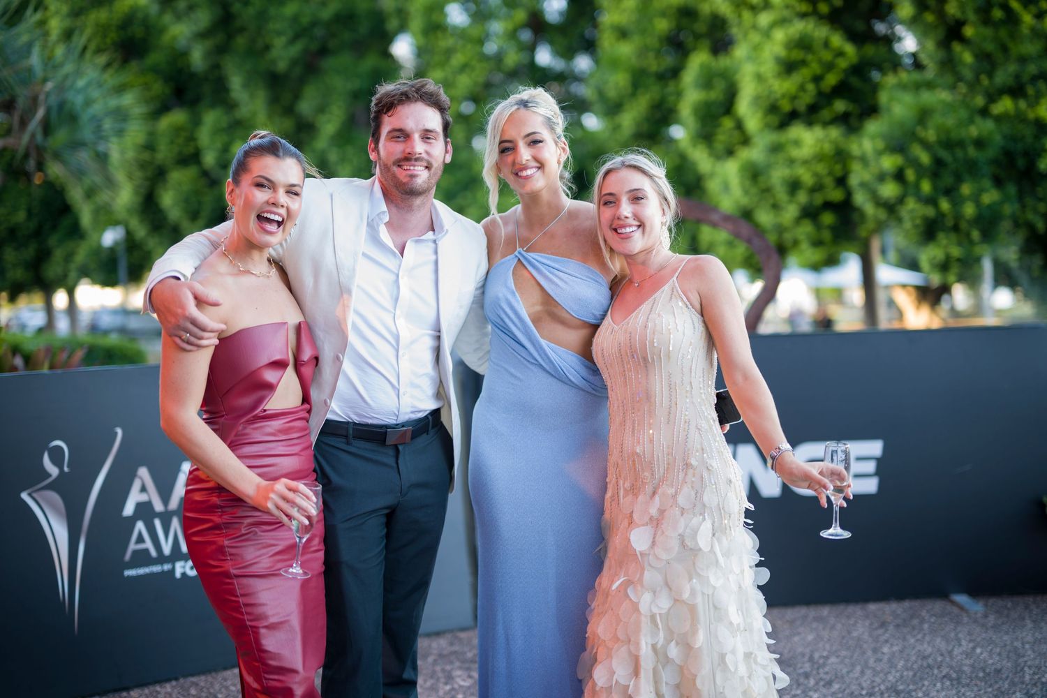 Four guests in colorful formal wear pose together at an outdoor event.