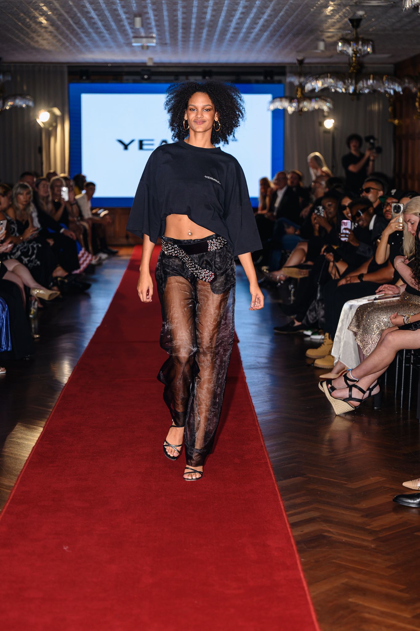 Model in black outfit with sheer lace pants walks runway at evening fashion event.