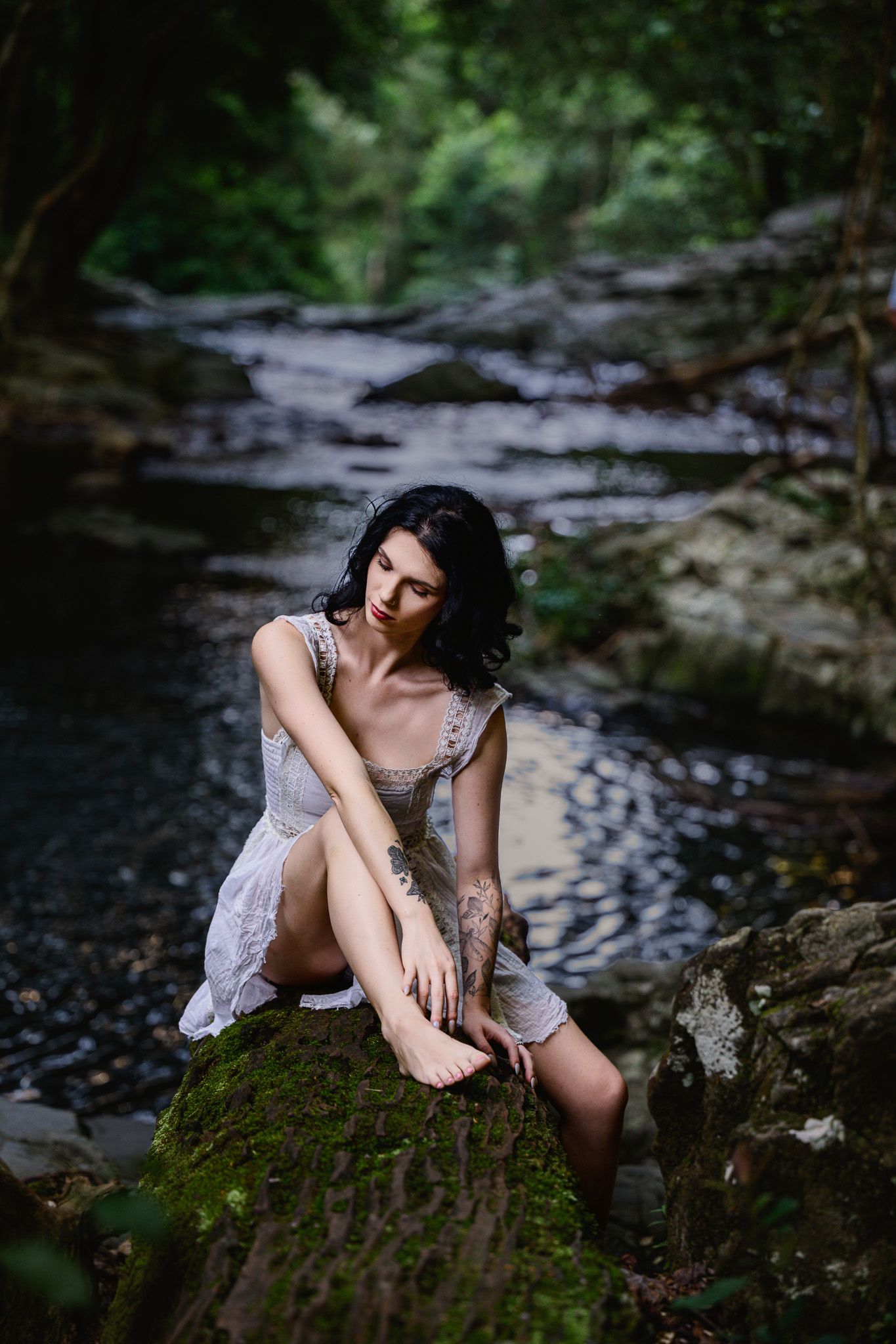 Woman in white lace dress sitting on mossy rock by forest stream, looking down pensively.