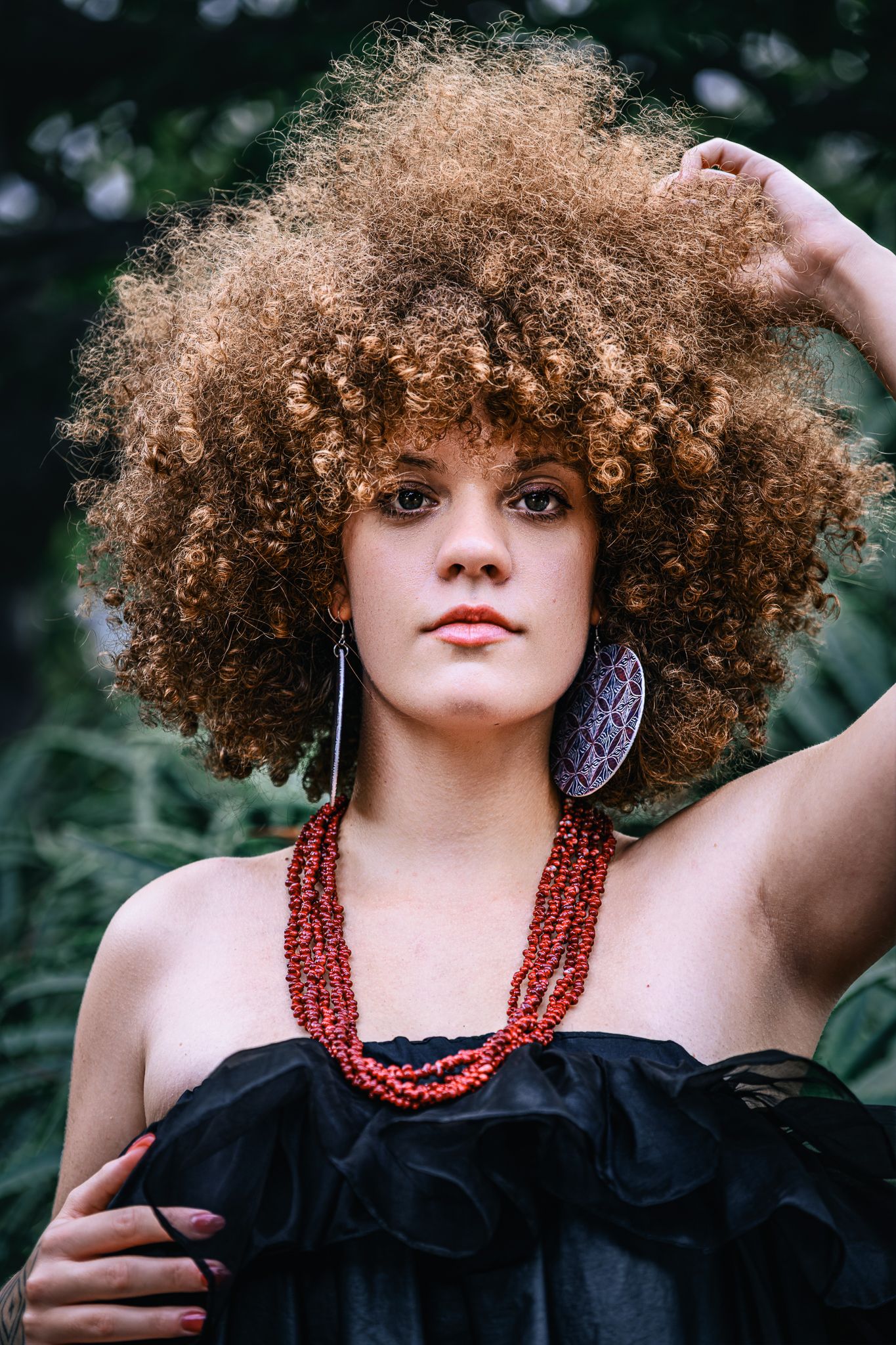 Woman with voluminous curly auburn hair wearing a black off-shoulder top, red beaded necklace, and large earrings outdoors.