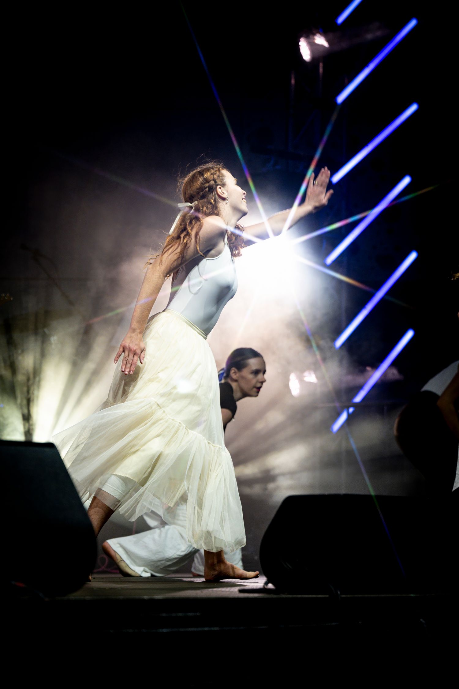 Performer in flowing white dress dancing dramatically on stage with blue lighting effects.