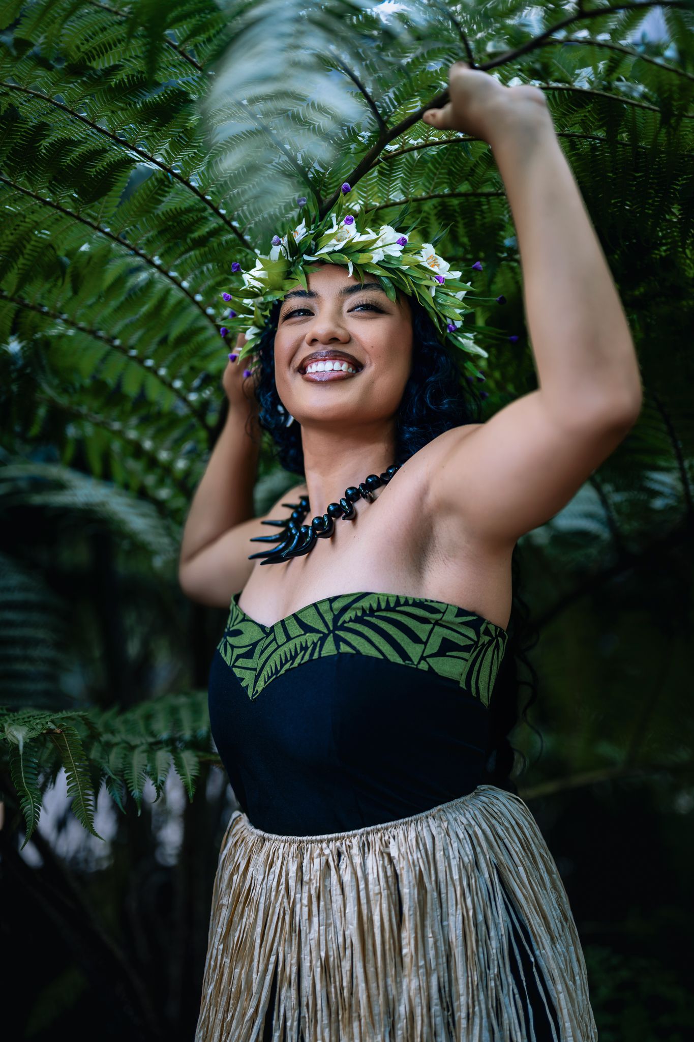 Smiling Hawaiian hula dancer in traditional grass skirt and floral lei headpiece posing among lush tropical ferns.