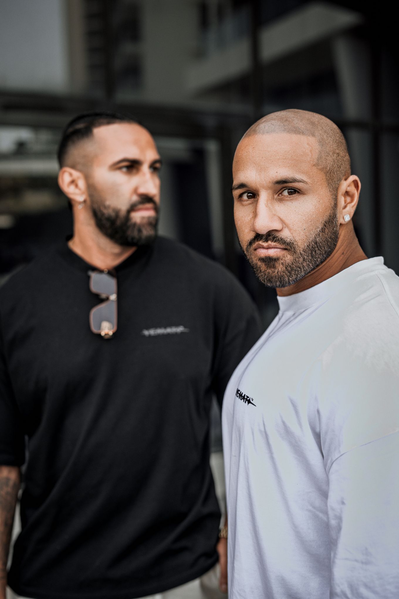 Two muscular bearded men in black and white t-shirts posing outdoors, one bald looking at camera.
