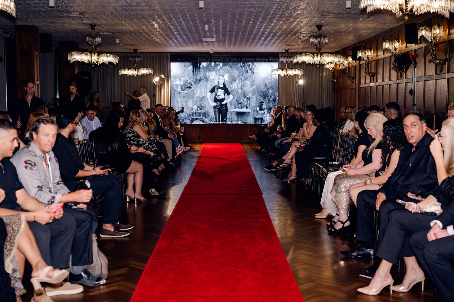 Audience members seated along a red carpet runway in an elegant indoor venue.