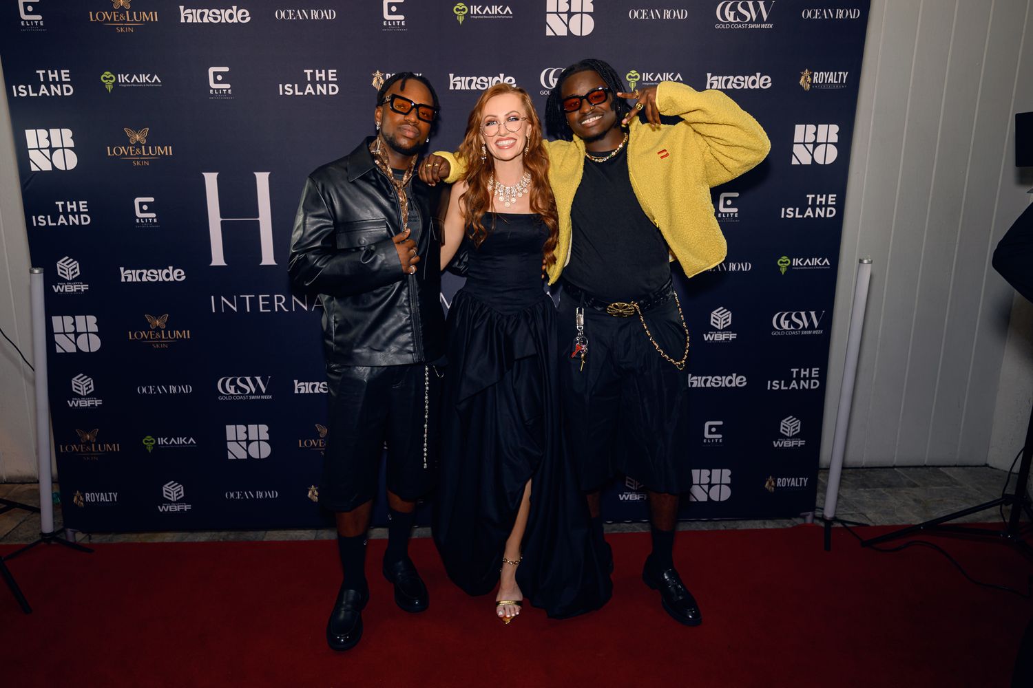 Three people pose together at a formal event against a branded backdrop on the red carpet.