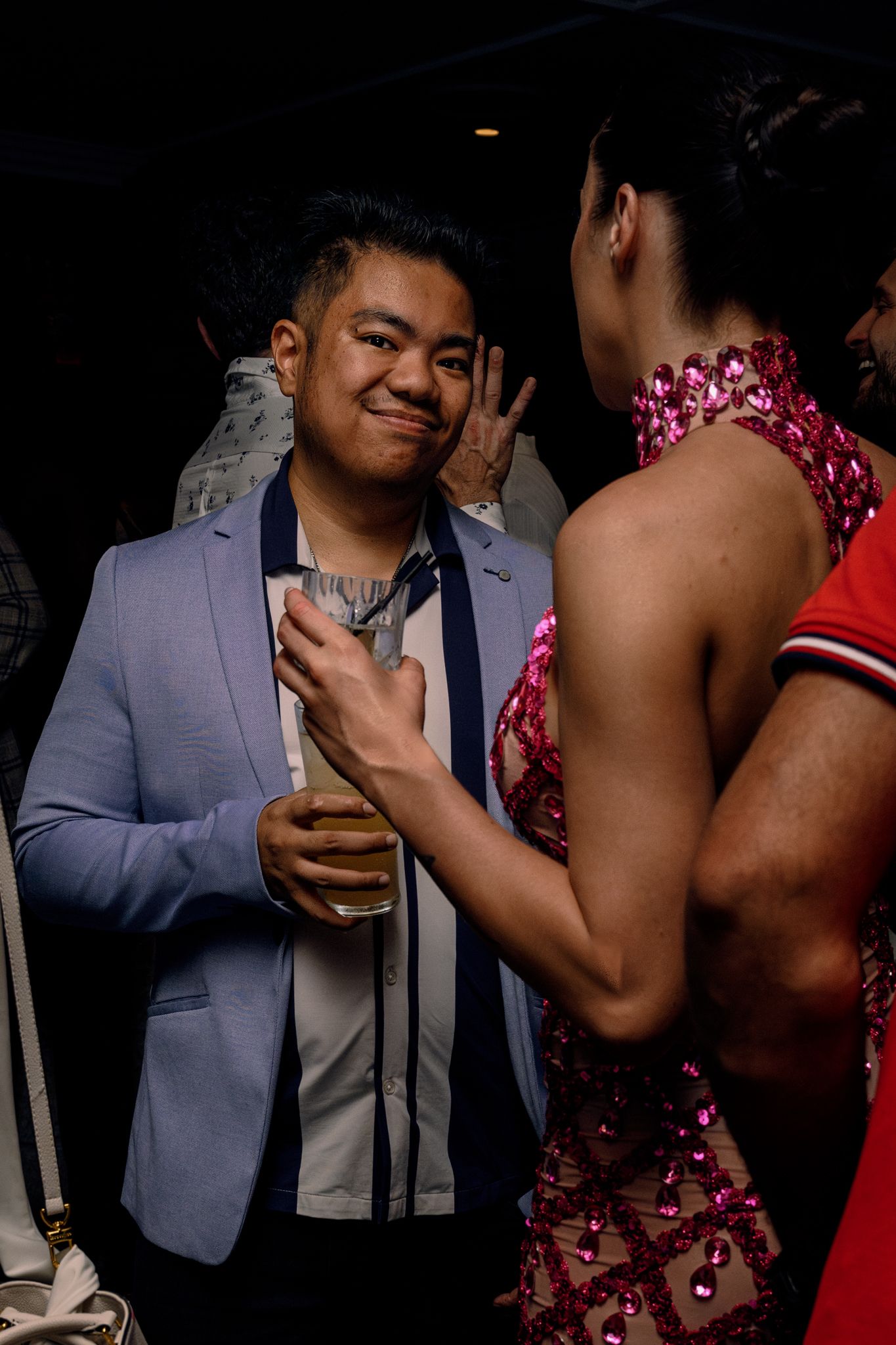 Man in light blue blazer holding drink smiles at person in pink sequin dress at a party.