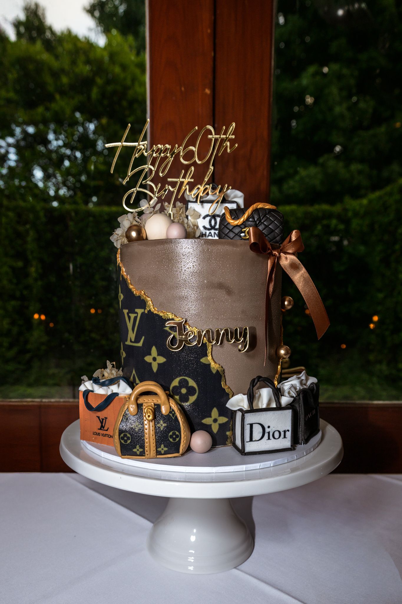 Designer cake decorated with luxury brand logos and metallic accents on white cake stand.