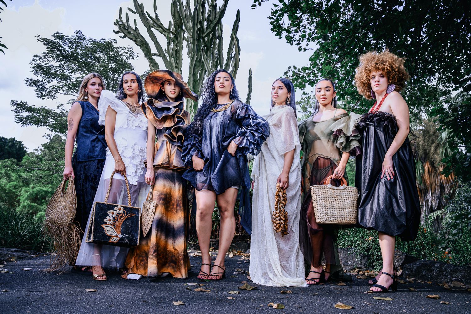 Seven diverse women pose outdoors in elegant fashion designs, surrounded by lush tropical greenery.