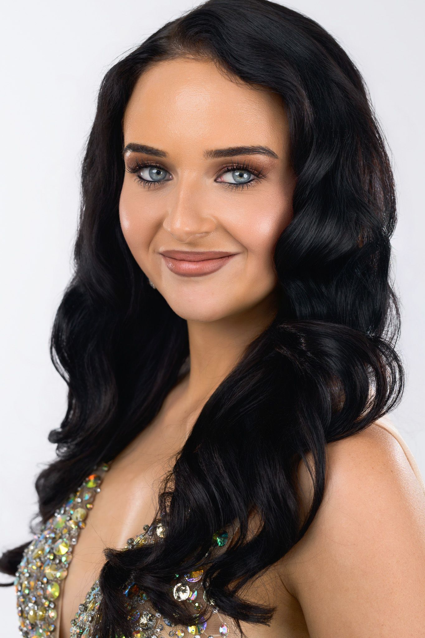 Young woman with long black wavy hair and blue eyes wearing a sequined dress, smiling against white background.