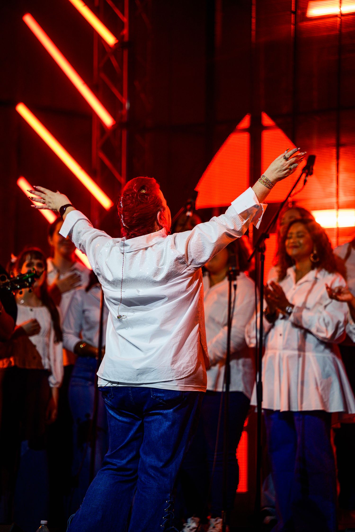 Worship leader in white shirt and blue pants leading praise with arms raised under red stage lighting.