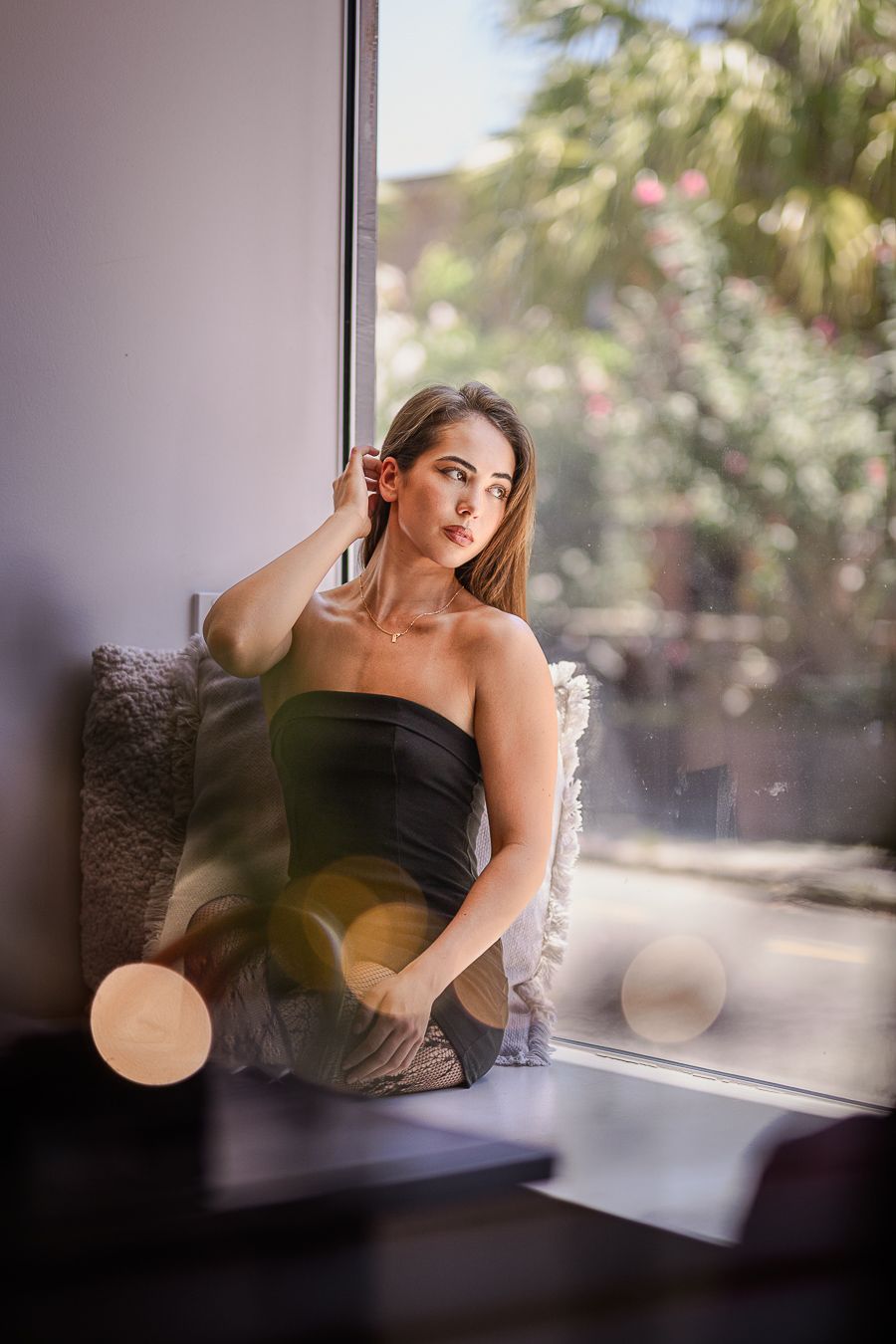 Person in black dress poses by window with palm trees visible outside.
