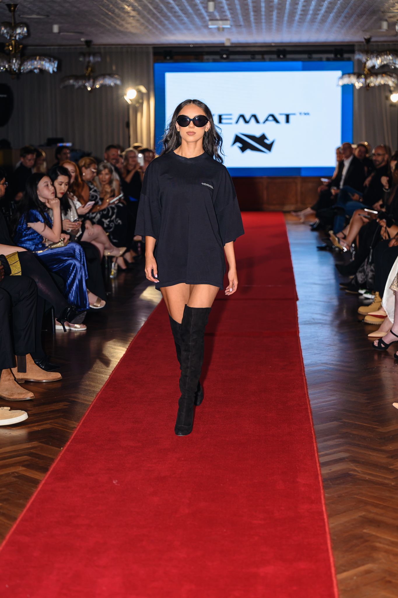 Model in all-black ensemble and sunglasses walks fashion runway with audience seated on both sides.