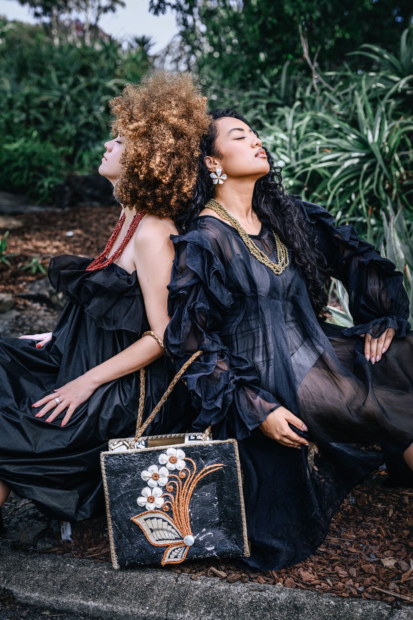 Two women in elegant black gowns pose outdoors with a decorative floral handbag, surrounded by lush greenery.
