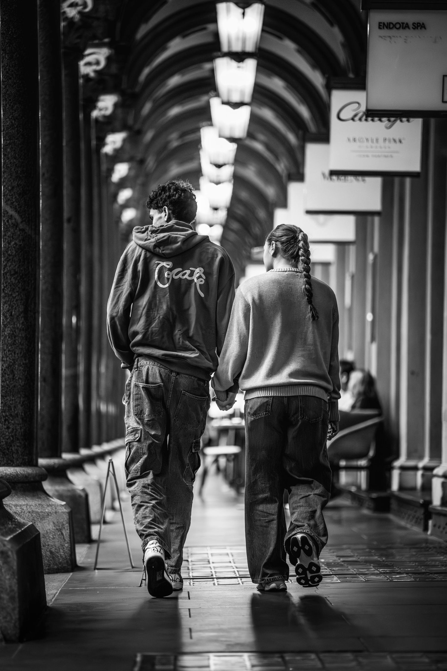 Black and white photo of a couple holding hands walking through an elegant covered arcade with arched ceilings.