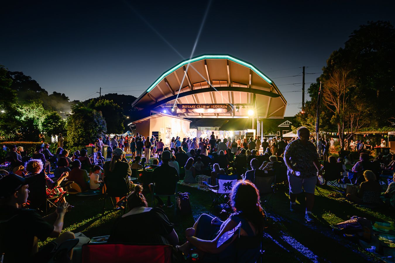 Crowd gathered at night for an outdoor concert at an illuminated amphitheater stage in a park.