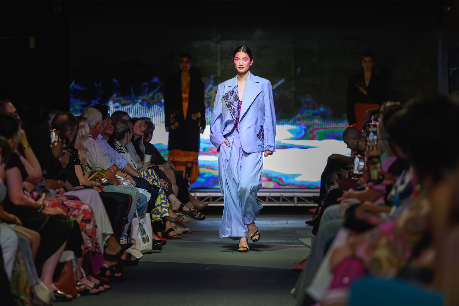 Model walks runway in light blue pantsuit at fashion show, audience seated on both sides watching.