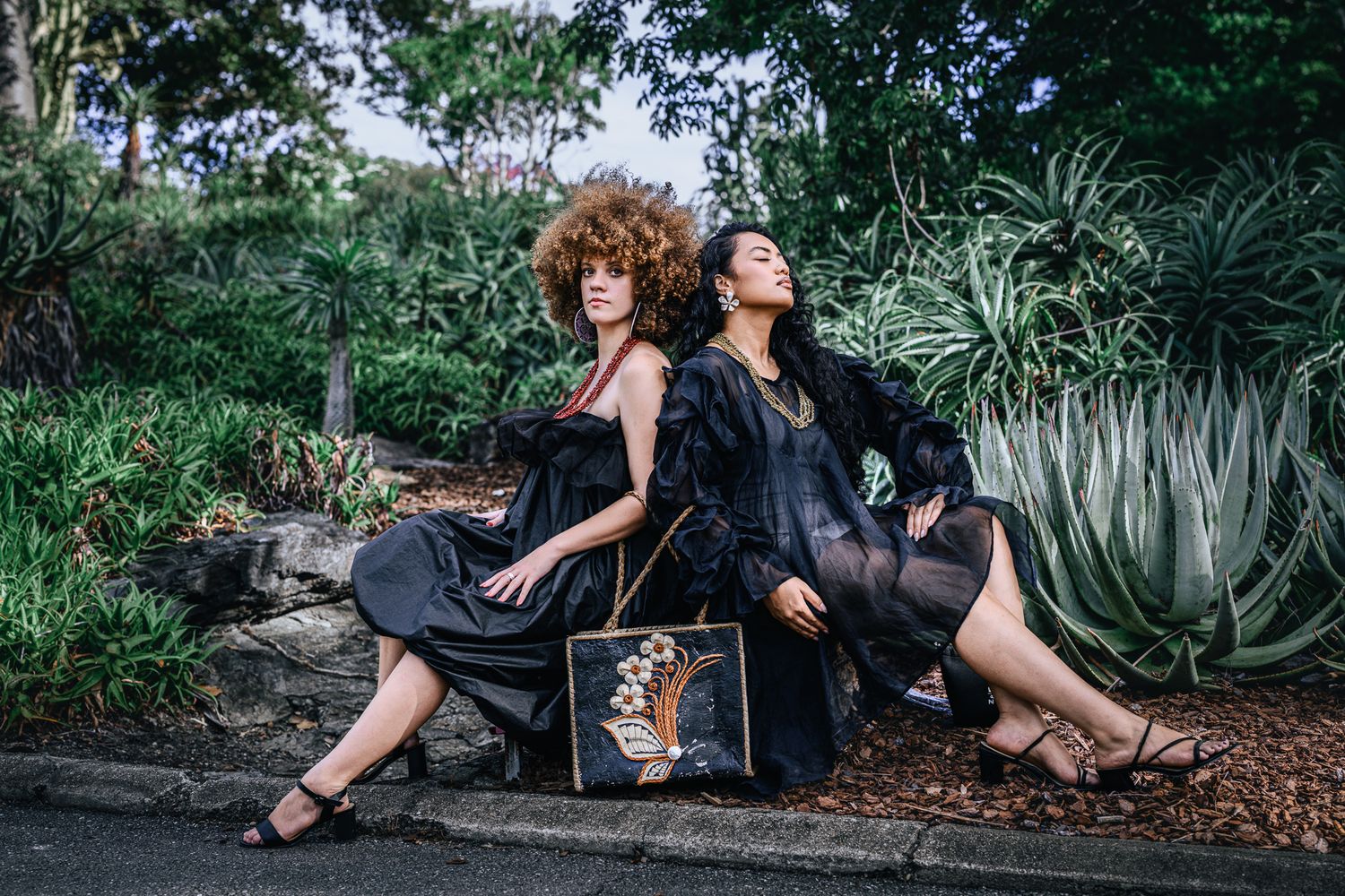 Two women in black dresses pose back-to-back outdoors with a decorative handbag among lush green plants.