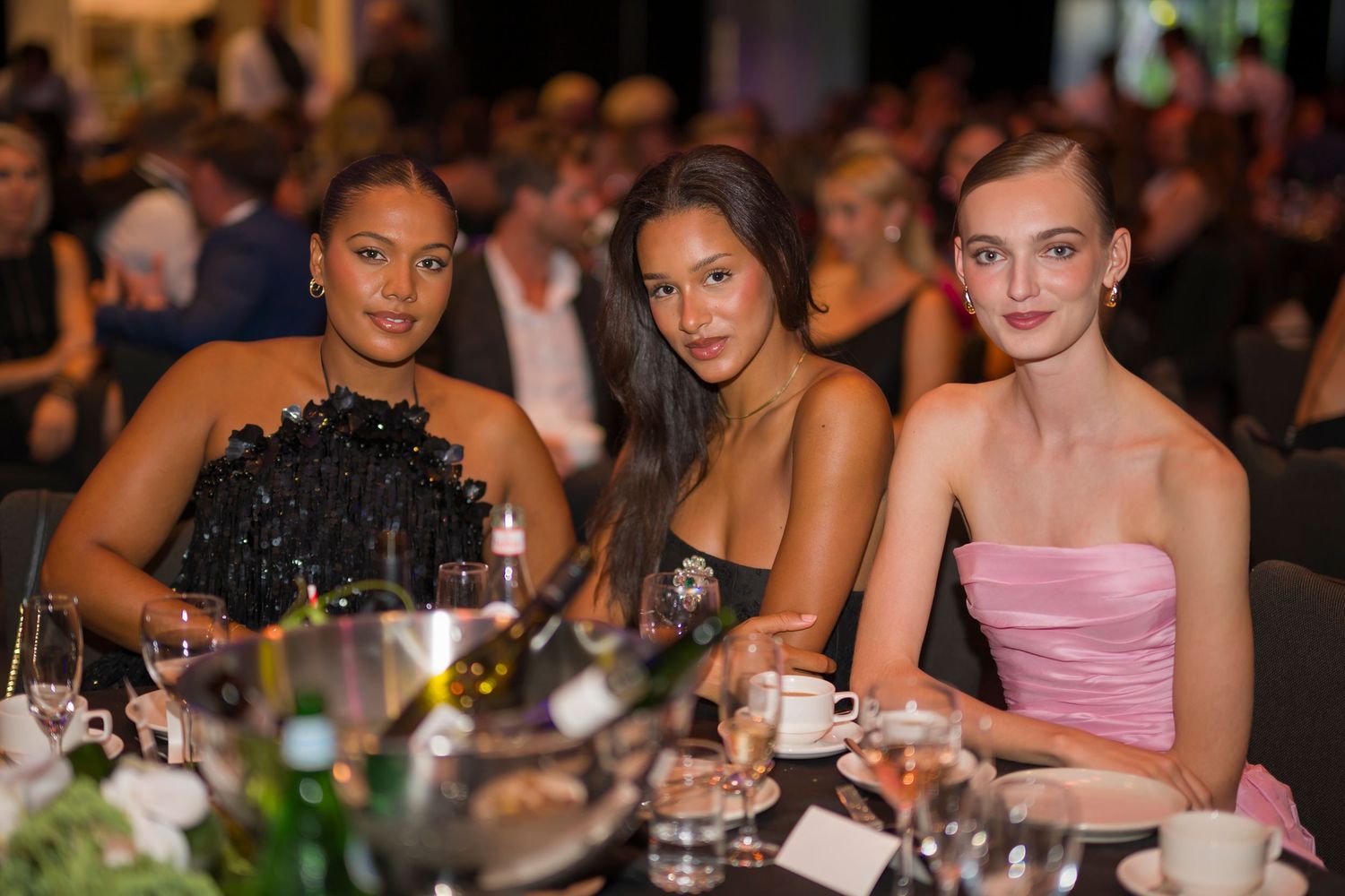 Three guests in elegant evening wear seated at formal dinner table.