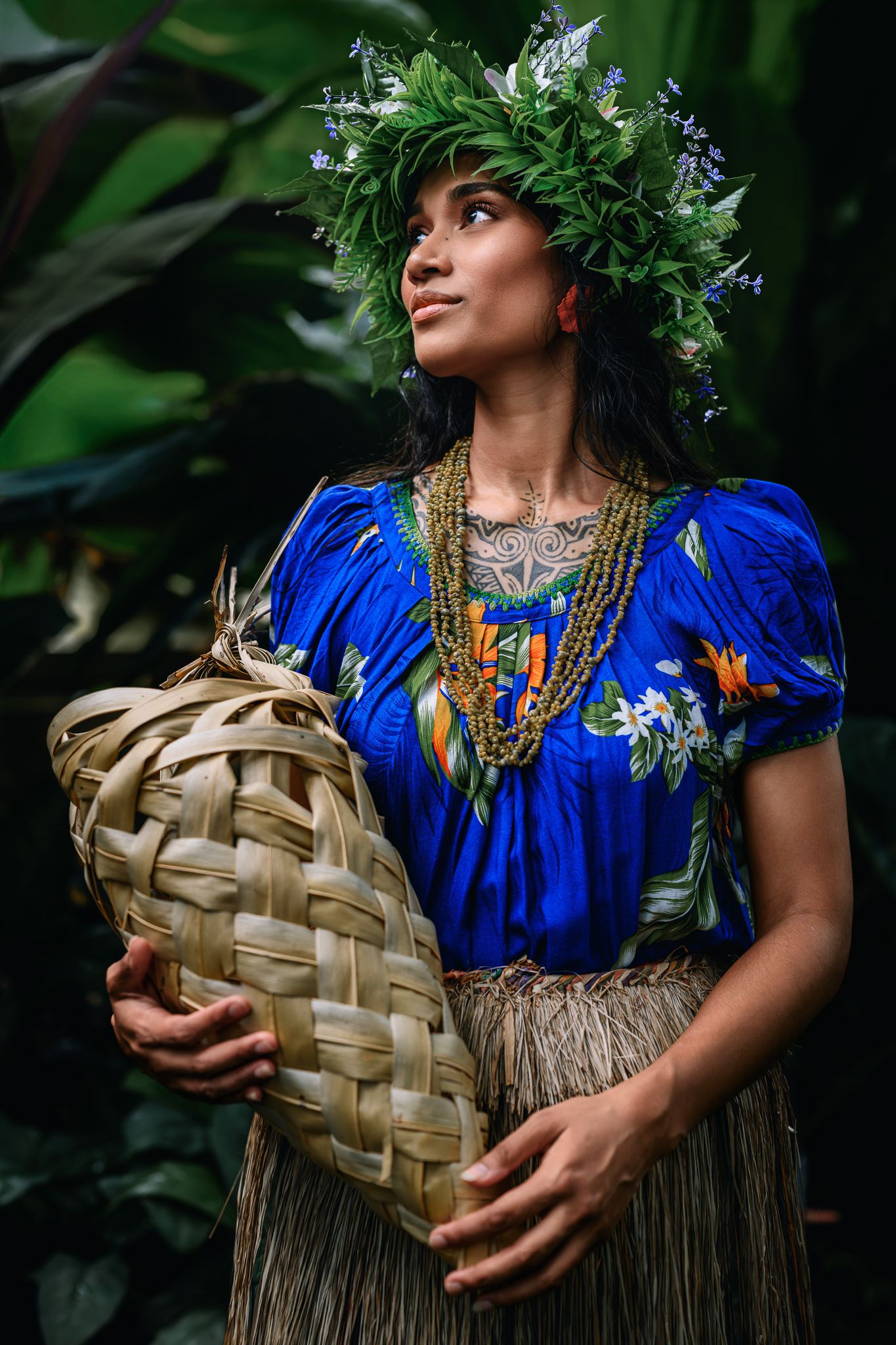 Woman in blue floral top, leaf crown, grass skirt, and beaded necklace holds woven basket amid tropical greenery.