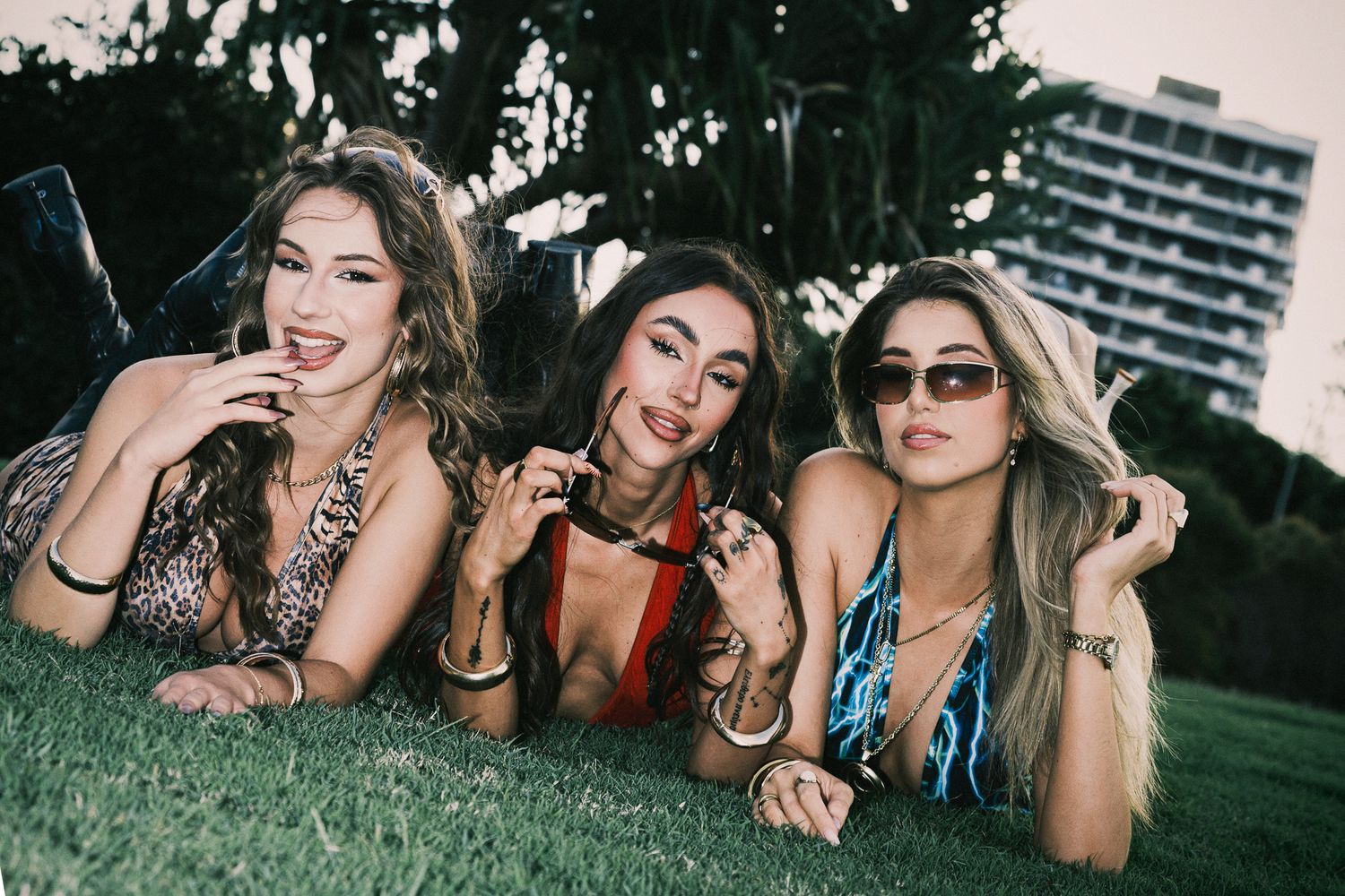 Three women in swimwear lying on grass, smiling and posing together outdoors near a building.