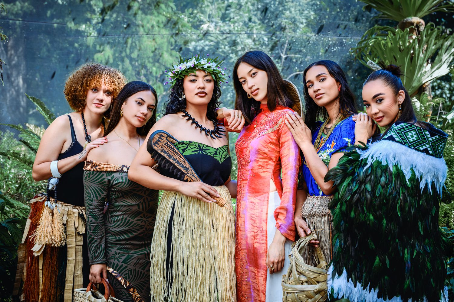 Six diverse women in traditional cultural attire pose together in a lush tropical garden setting.