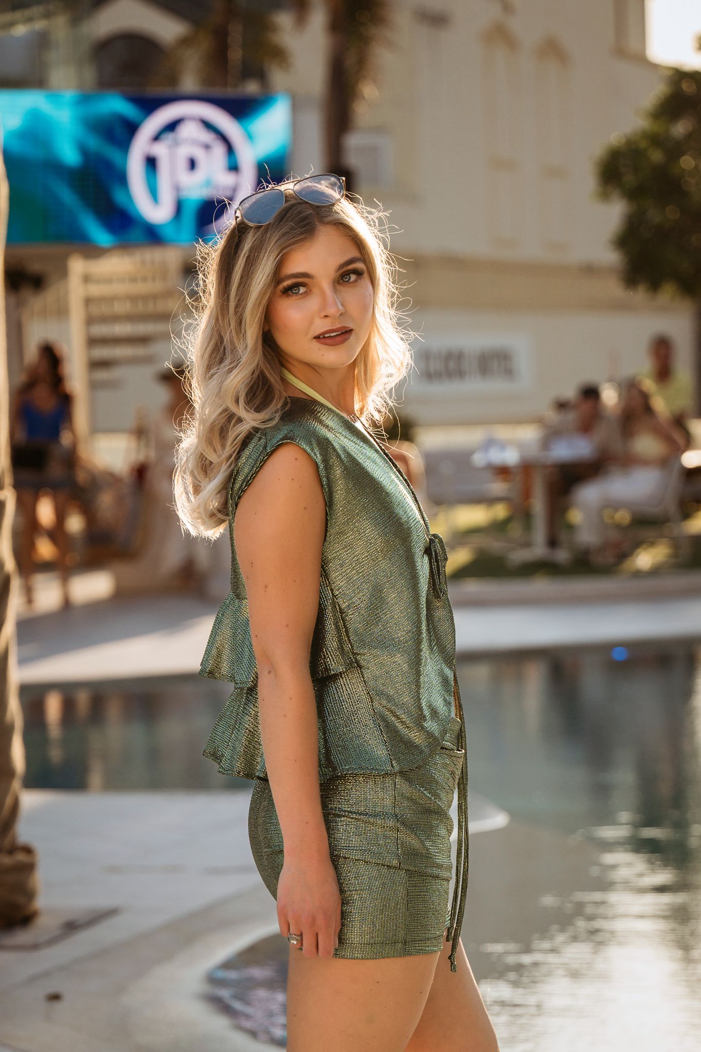 Model in sage green romper poses by pool during outdoor fashion event.