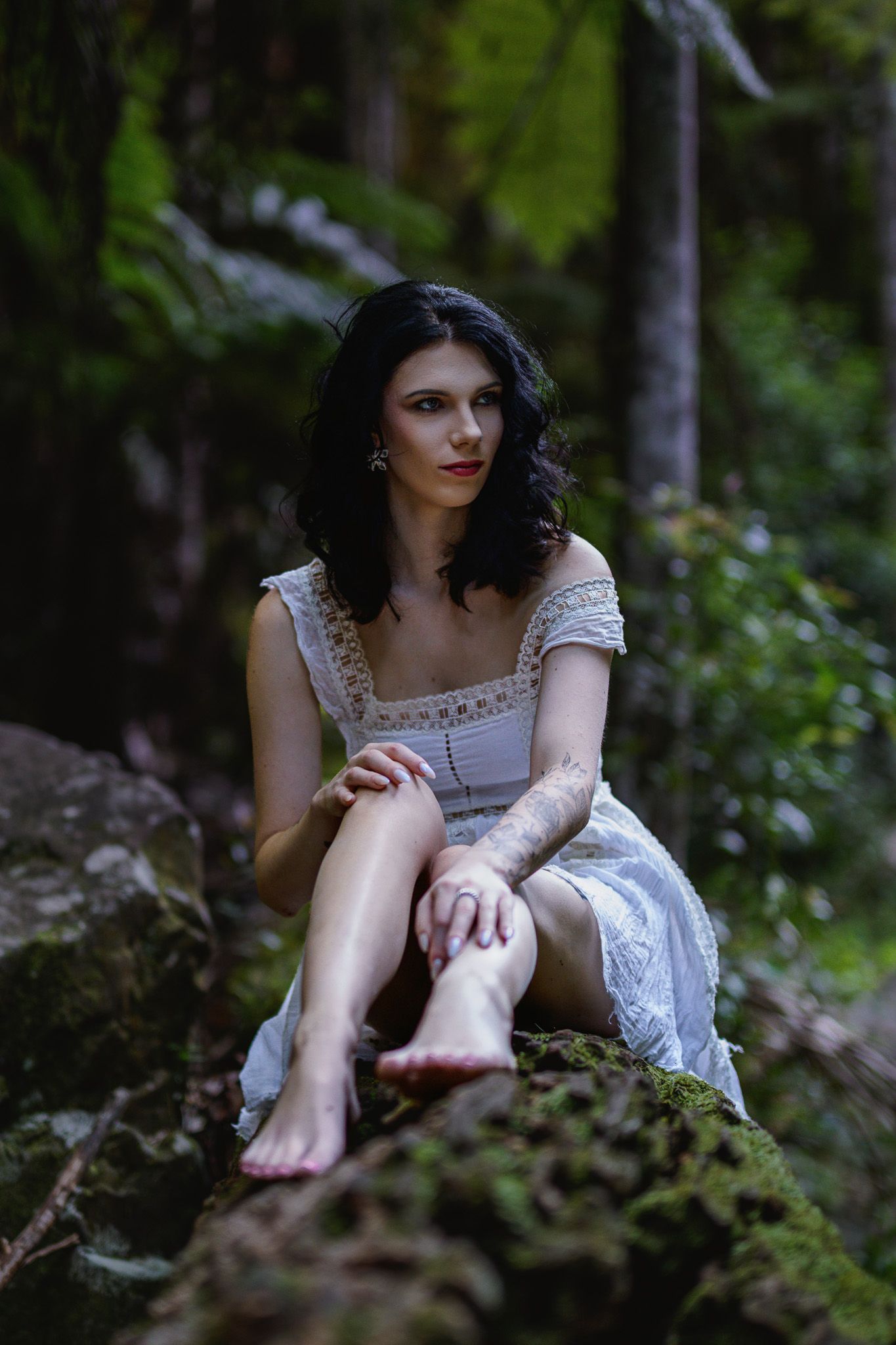 A barefoot woman in a white dress sits on a mossy rock in a lush green forest, gazing thoughtfully into the distance.