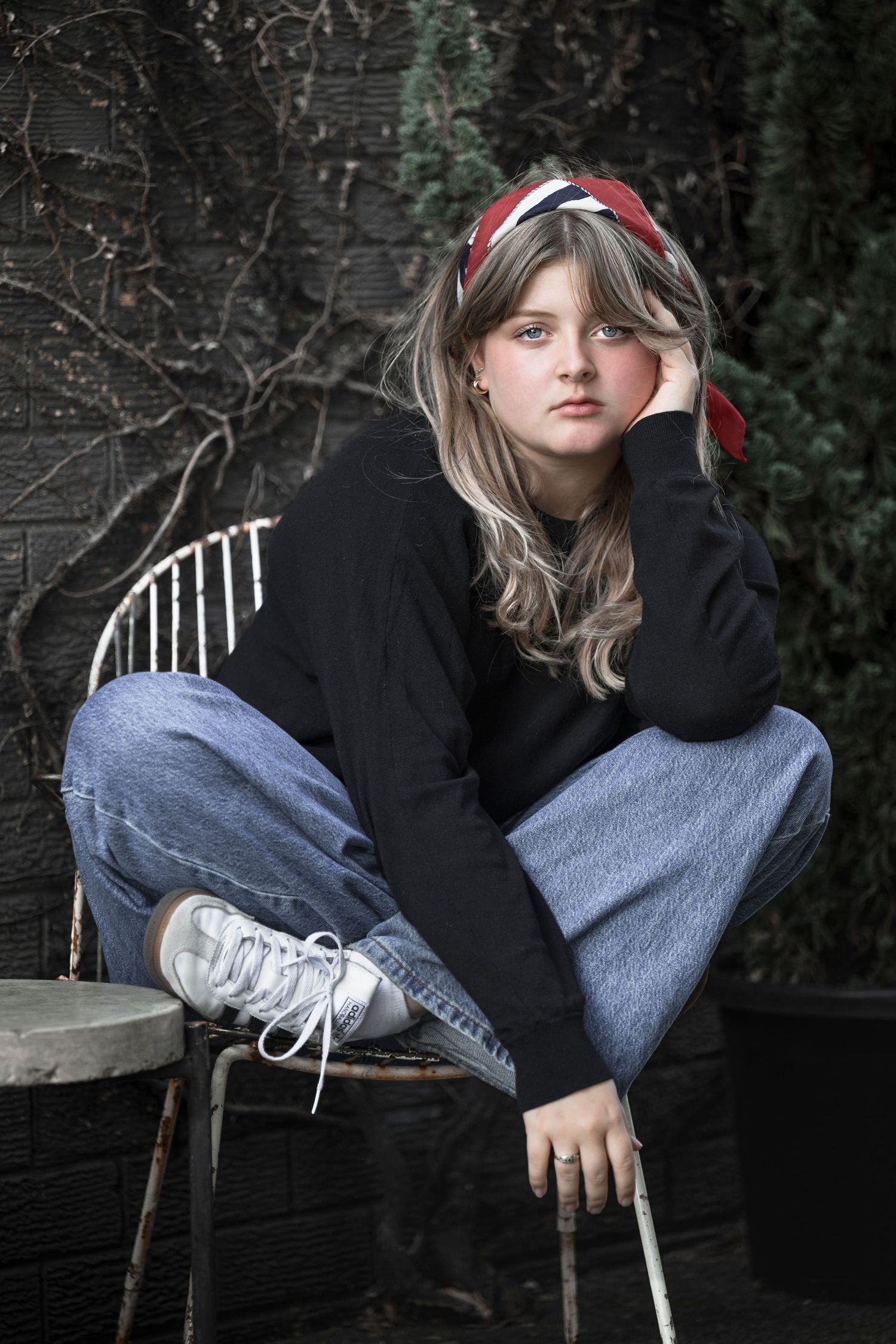 Young girl in black sweater, jeans, and red headband sits cross-legged on a chair outdoors.