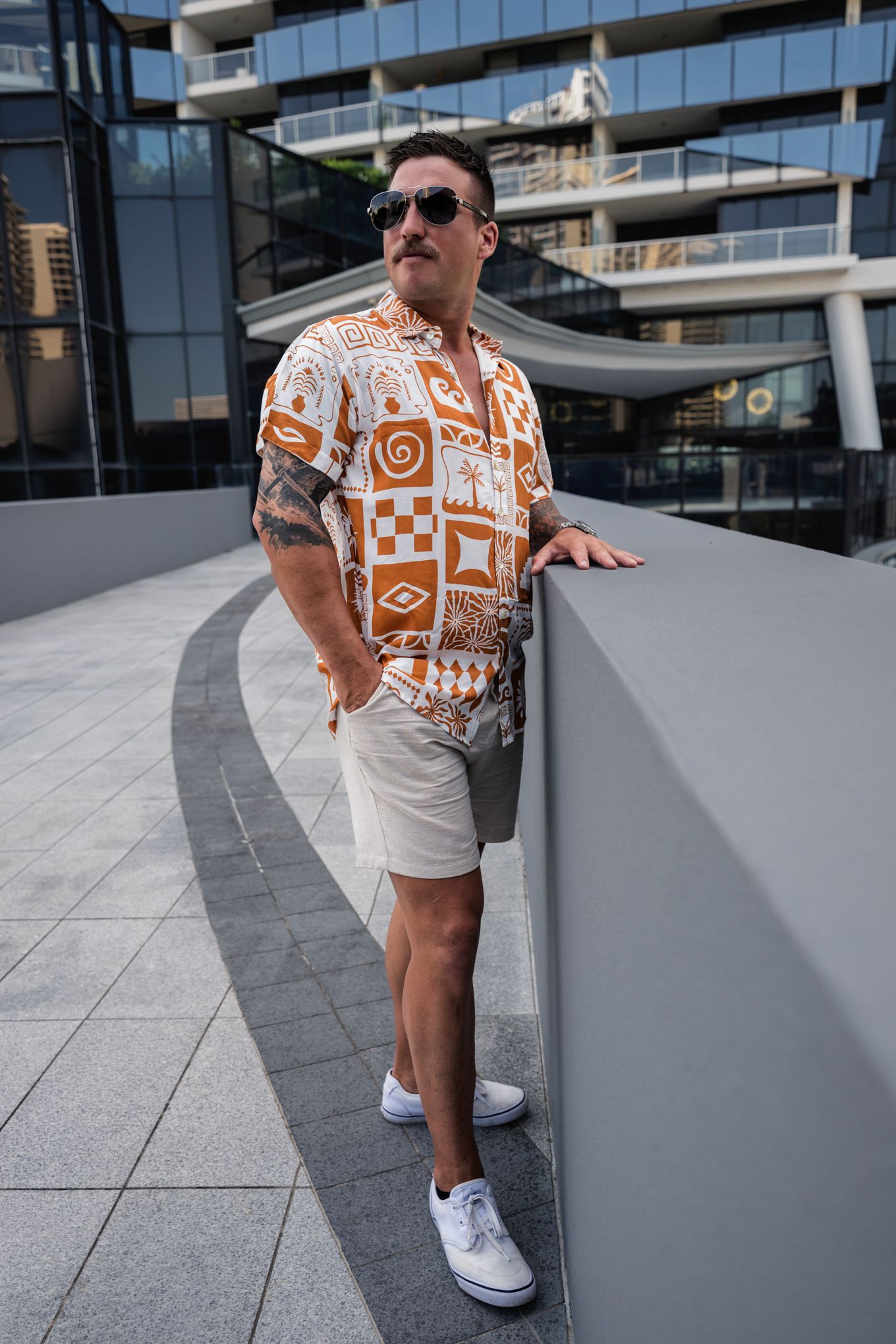 Person in geometric print shirt and white shorts leaning against modern building wall.