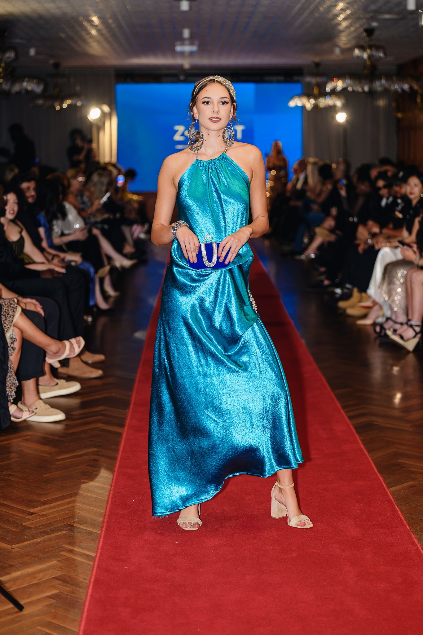 Model in turquoise silk gown poses dramatically on red carpet runway.