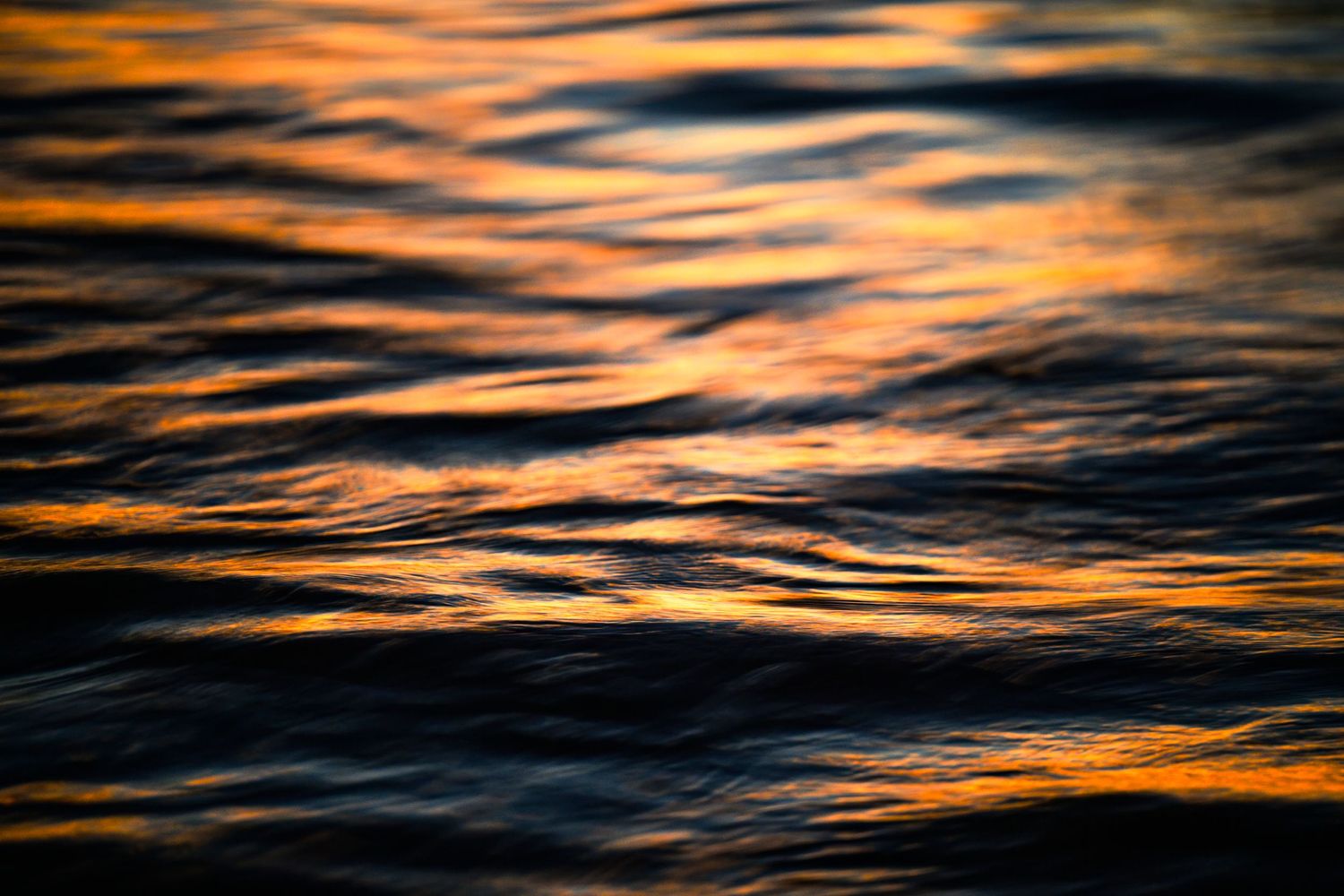 Golden sunset reflections shimmer on dark ocean water waves creating an abstract pattern of orange and black hues.
