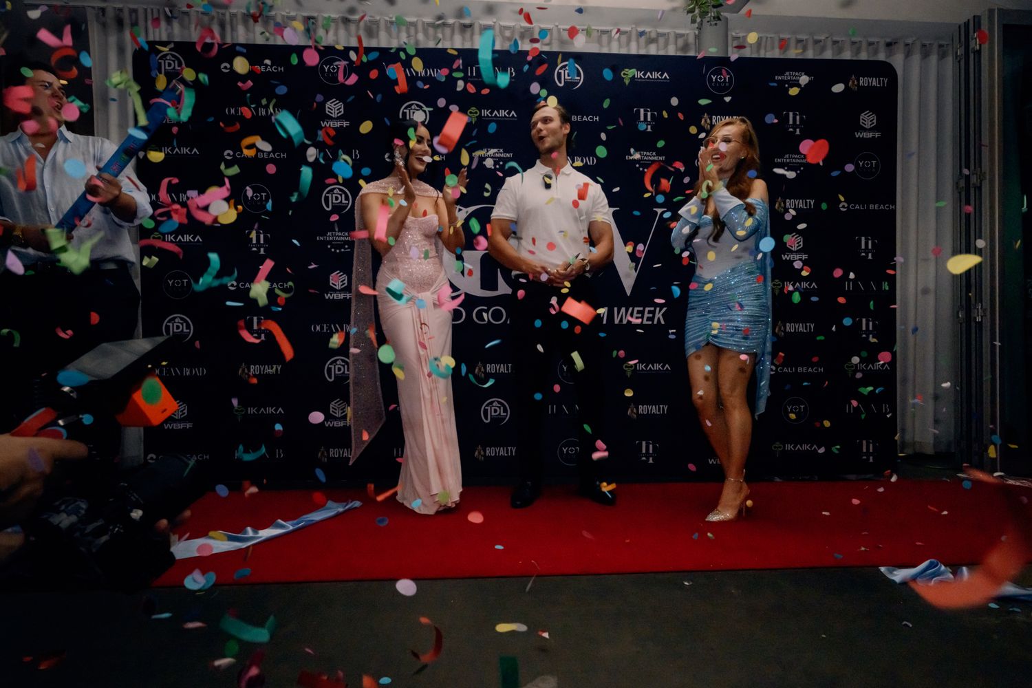 Three people celebrate on a red carpet with colorful confetti falling at a festive event.