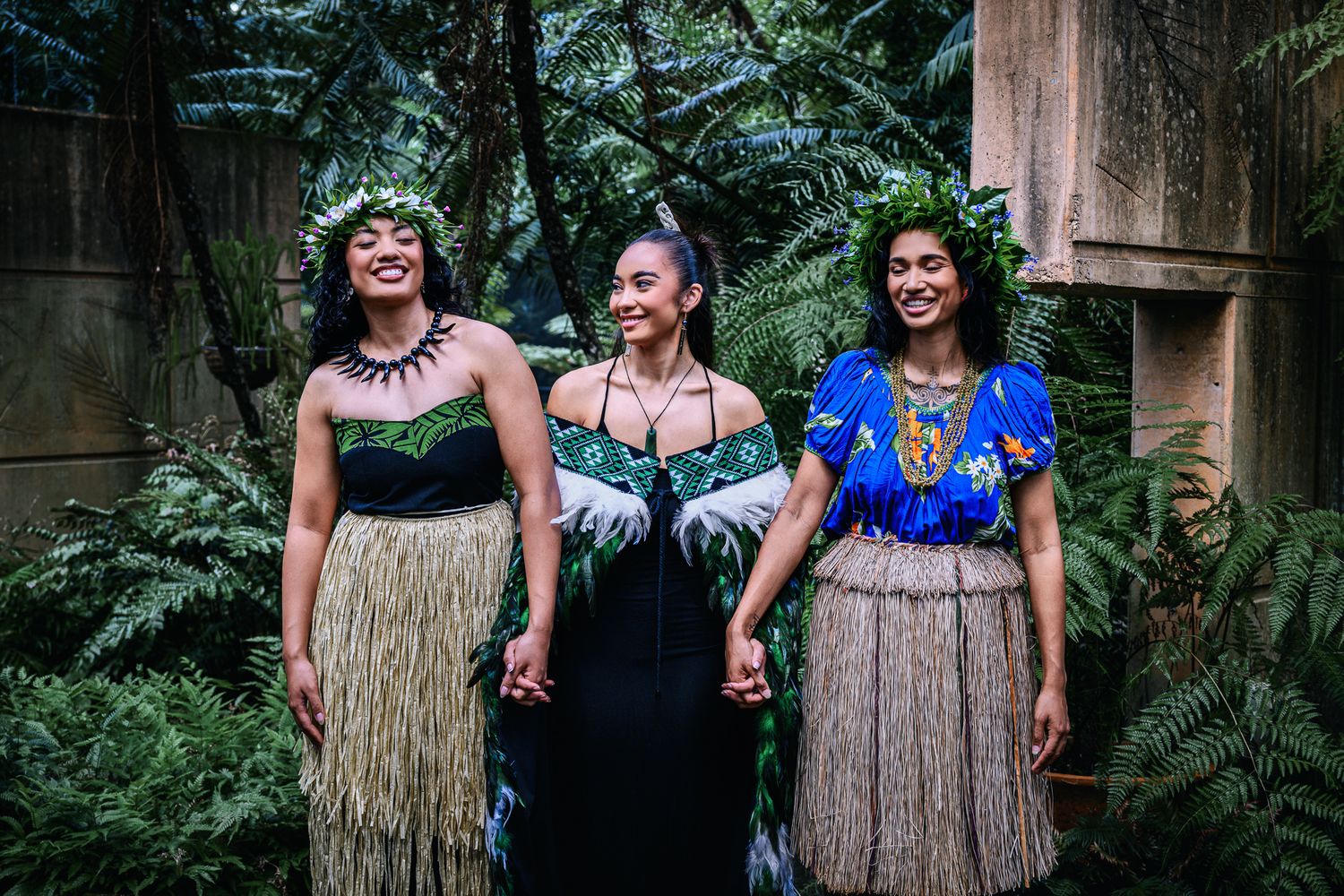 Three women in traditional Polynesian attire with grass skirts and floral headpieces smiling in a lush garden.