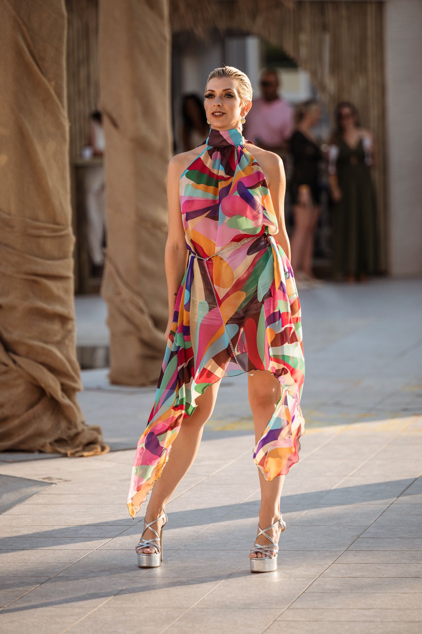 Model wearing colorful abstract print wrap dress poses on runway with platform heels.