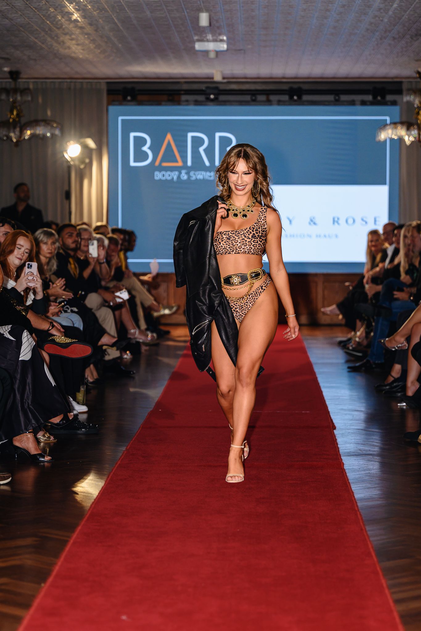 Model walks runway in revealing swimwear and black jacket at fashion show.