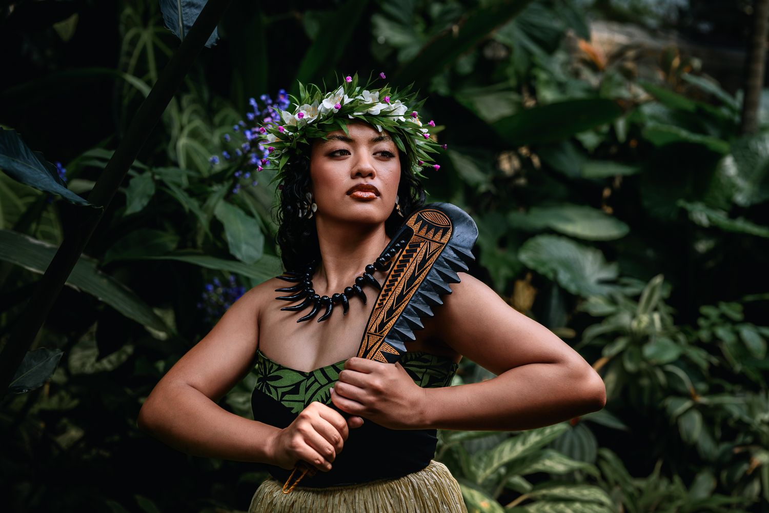 A Hawaiian hula dancer in traditional attire with floral crown and grass skirt poses gracefully among lush tropical foliage.