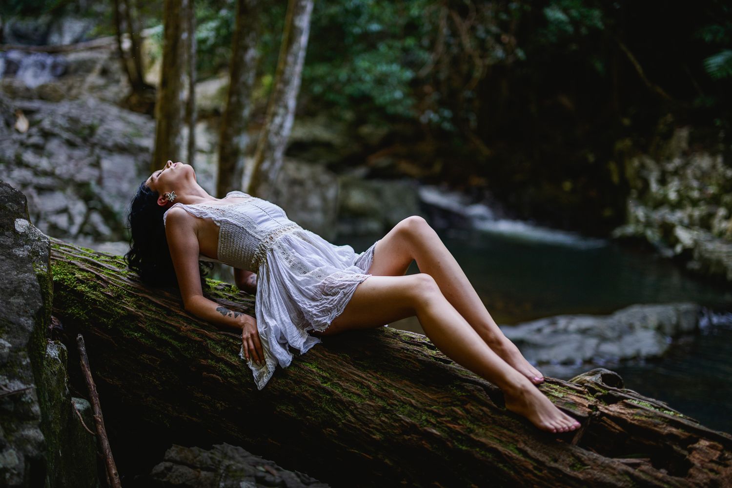 Woman in white dress reclining on mossy log in lush forest near stream.
