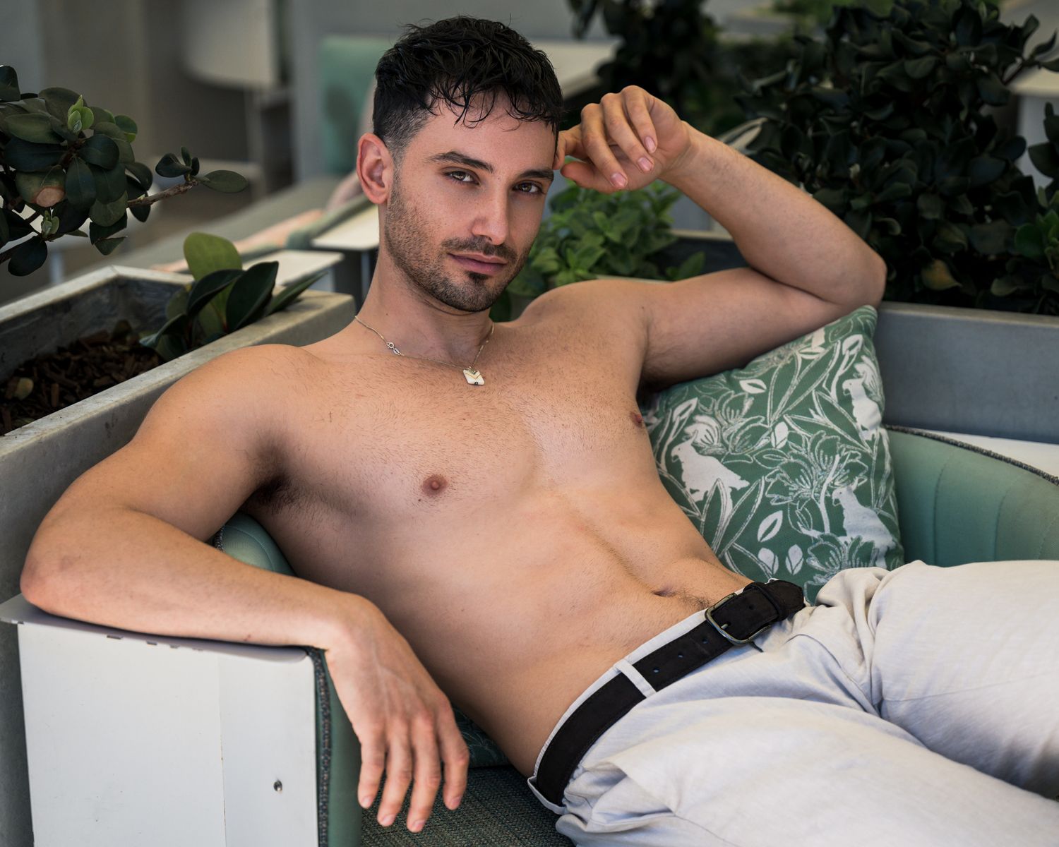 Shirtless man lounging on white outdoor furniture with tropical plants in background.