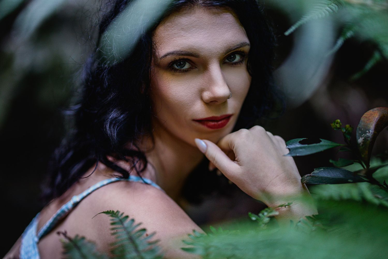 Dark-haired woman with red lips poses among lush green ferns and leaves in a moody forest portrait.
