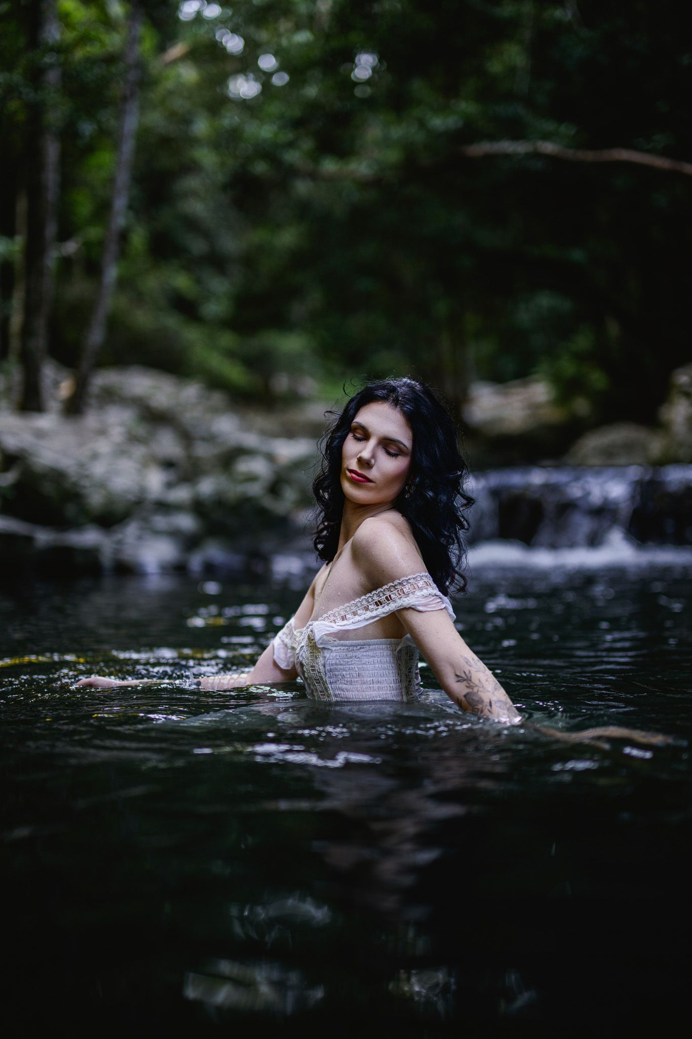 Dark-haired woman in white lace dress standing in a forest stream with waterfall backdrop.