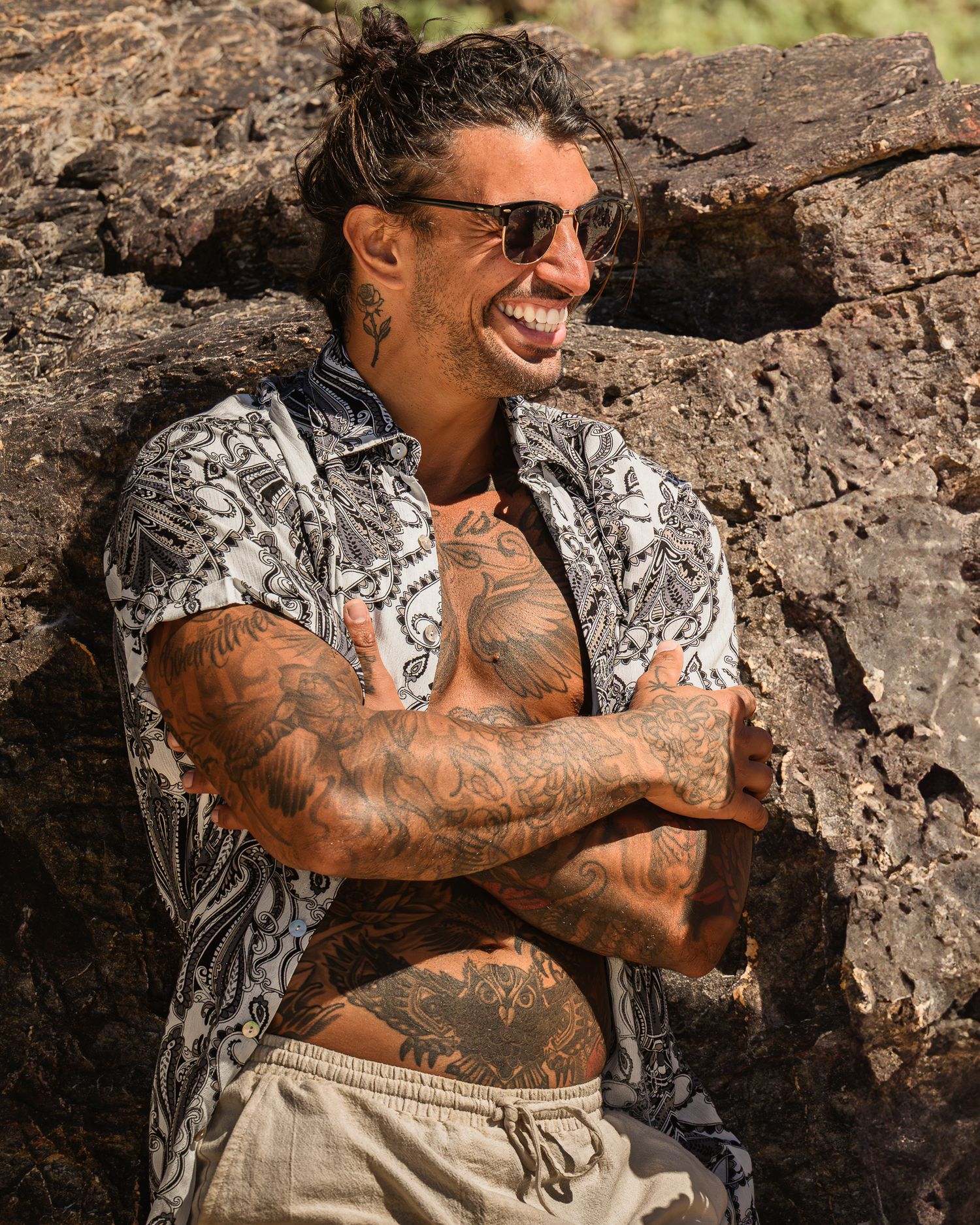 Tattooed model in patterned shirt smiles while posing against rocky backdrop.