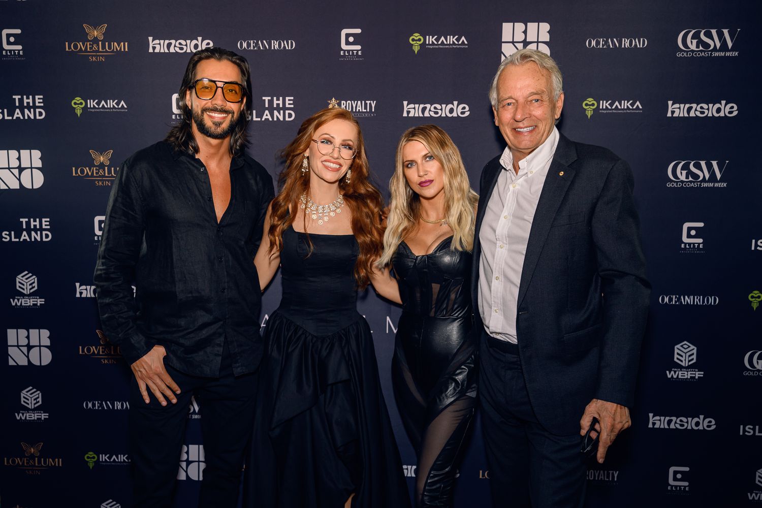 Four people in formal attire pose together at an upscale event with a branded backdrop.