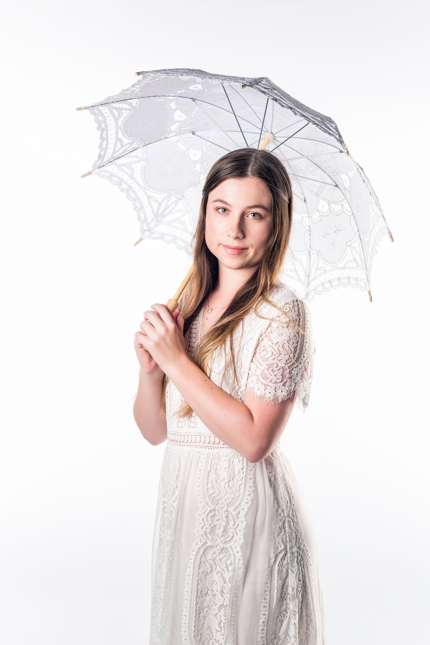 Model in white lace dress holds delicate white lace parasol against bright background.