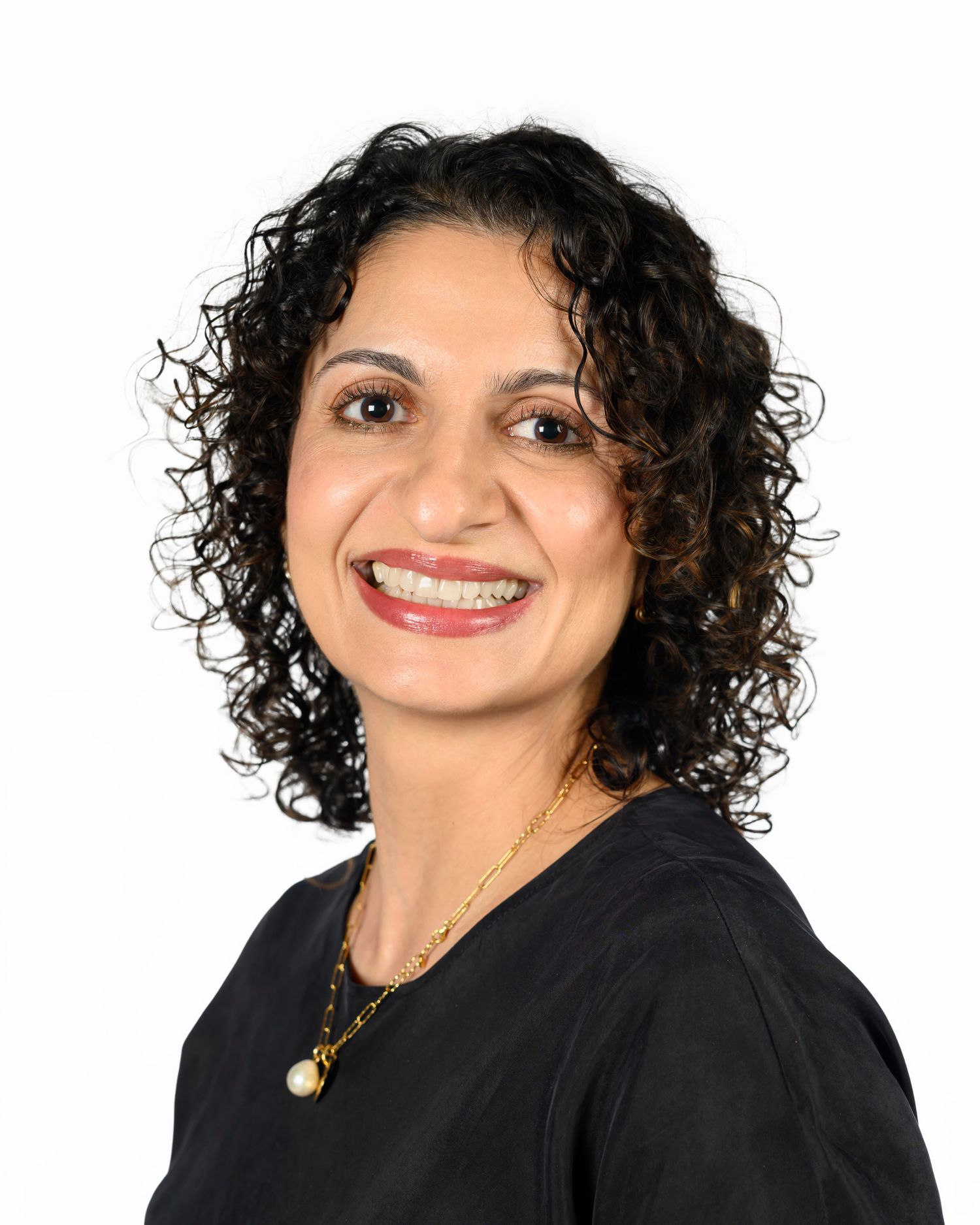 Professional headshot of a person with curly dark hair wearing a black top and necklace against a white background.