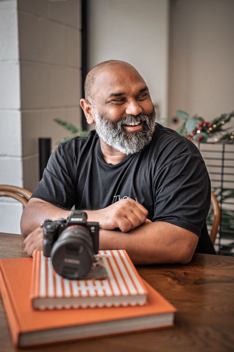 A photographer with a grey beard smiles while sitting at a wooden table with a camera and notebook.